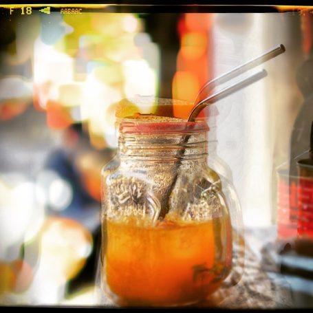A jar of orange drink with a straw, blurred colourful background.